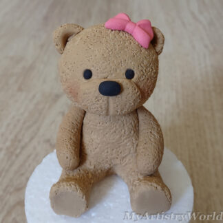 Handmade edible brown teddy bear cake topper with pink bow sitting on a white cake