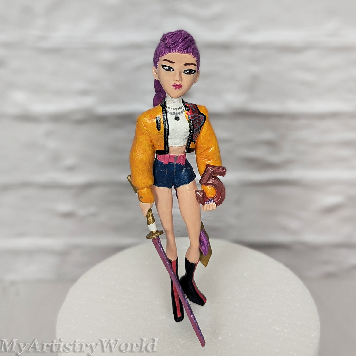 Handmade custom Rumi cake topper inspired by KPop Demon Hunters, sculpted from clay with purple hair, yellow jacket, sword, and personalized number